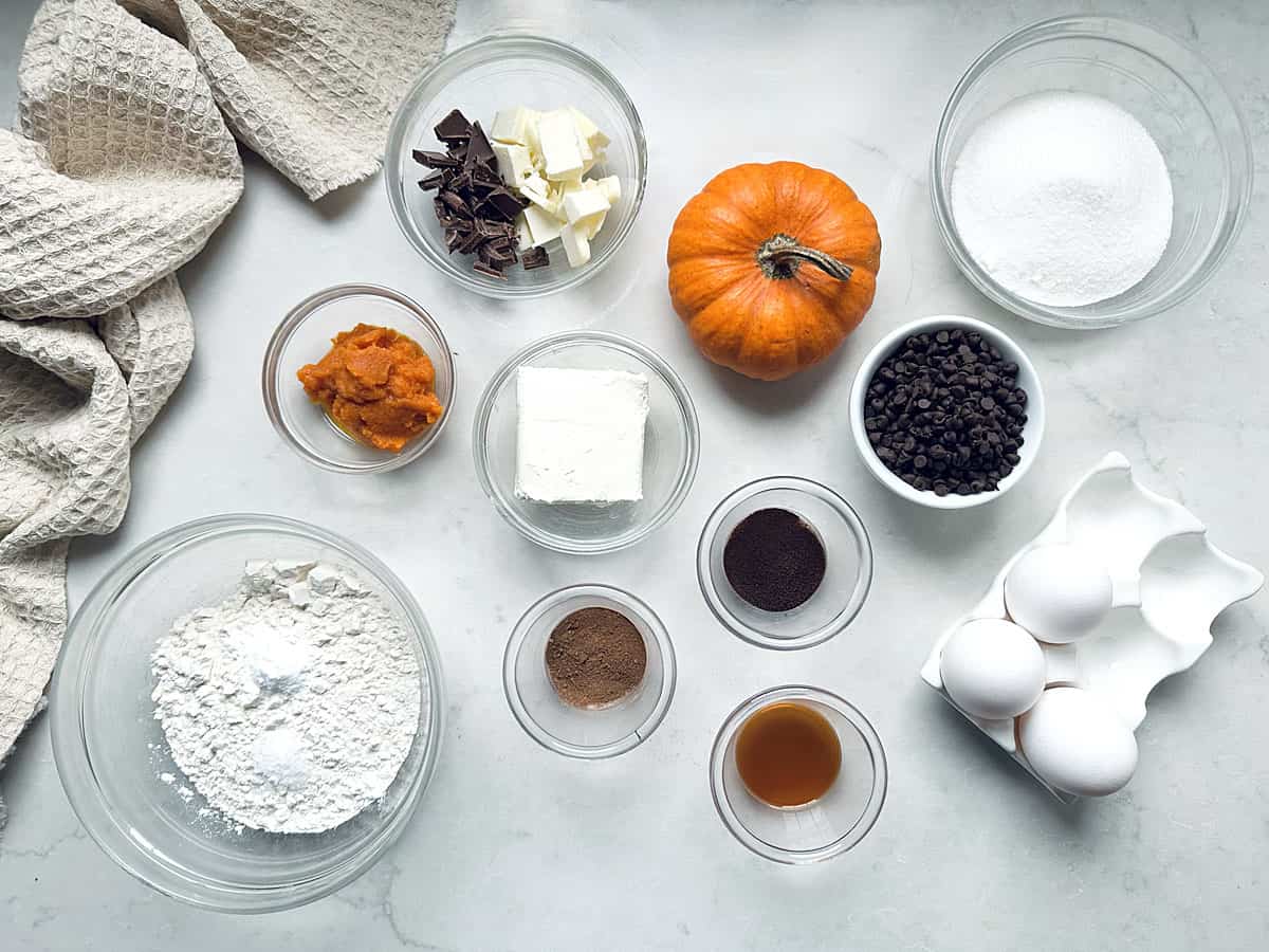 Baking ingredients arranged on a counter, including a small pumpkin, bowls of flour, sugar, chocolate chips, butter, pumpkin puree, cream cheese, spices, vanilla, and a carton of eggs.