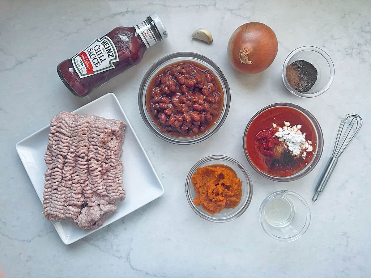Ingredients for a turkey pumpkin chili recipe arranged on a counter: ground meat, kidney beans, pumpkin puree, a bottle of Heinz chili sauce, an onion, a garlic clove, spices, and a small whisk.