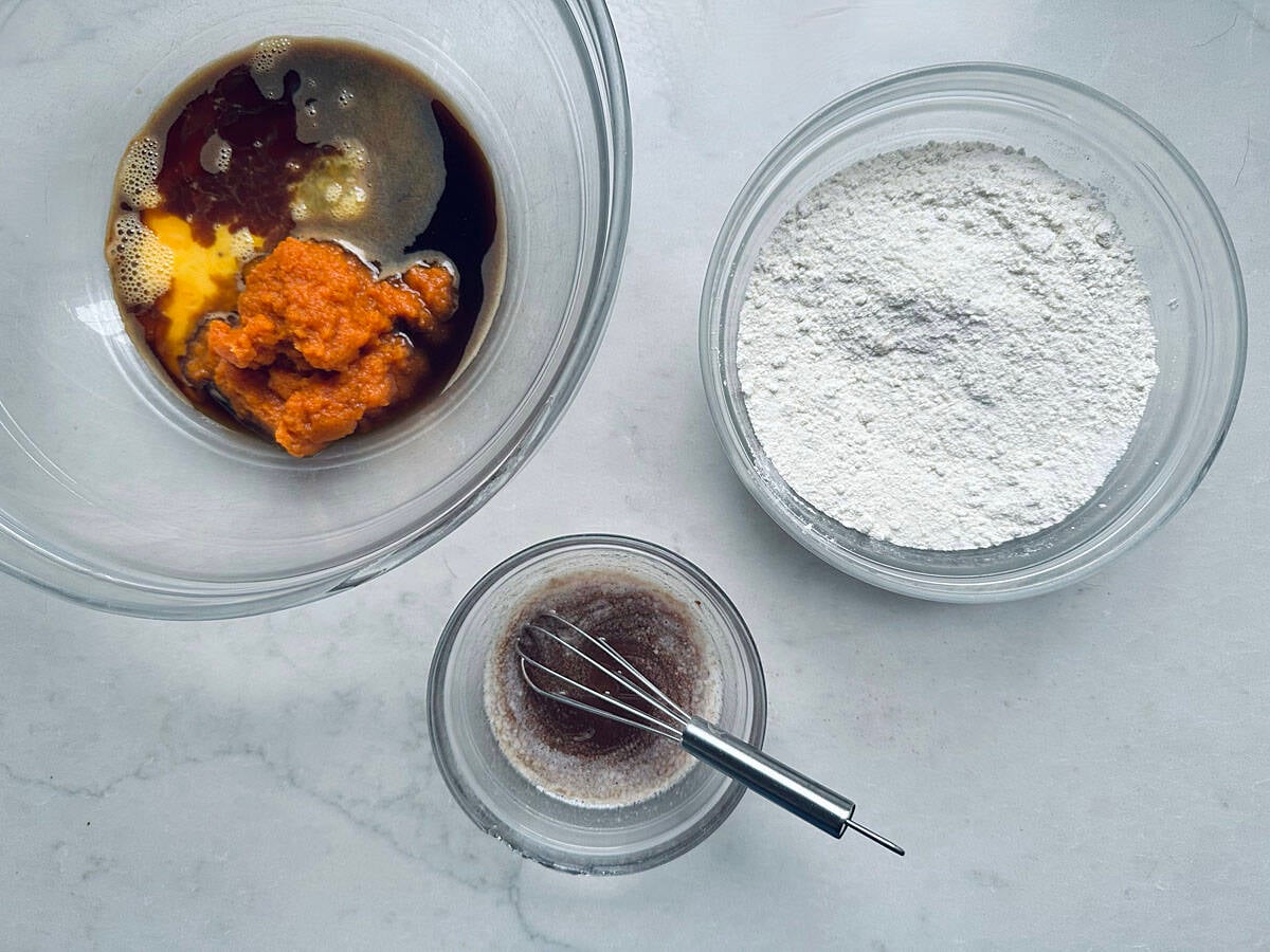 Three glass bowls containing pumpkin puree and liquid ingredients, dry flour, and a whisked mixture on a white surface.
