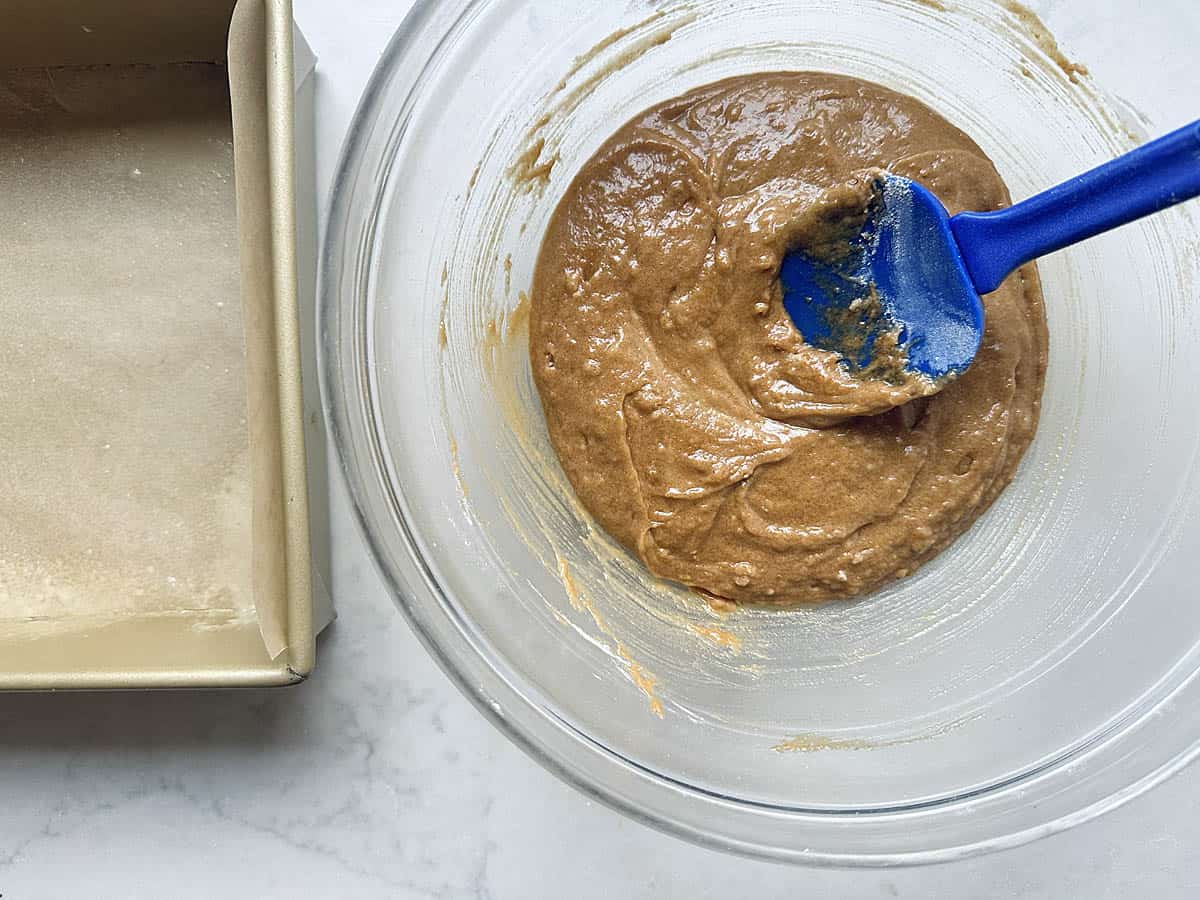 A blue silicone spatula mixes brown cake batter in a glass bowl next to a parchment-lined metal loaf pan.