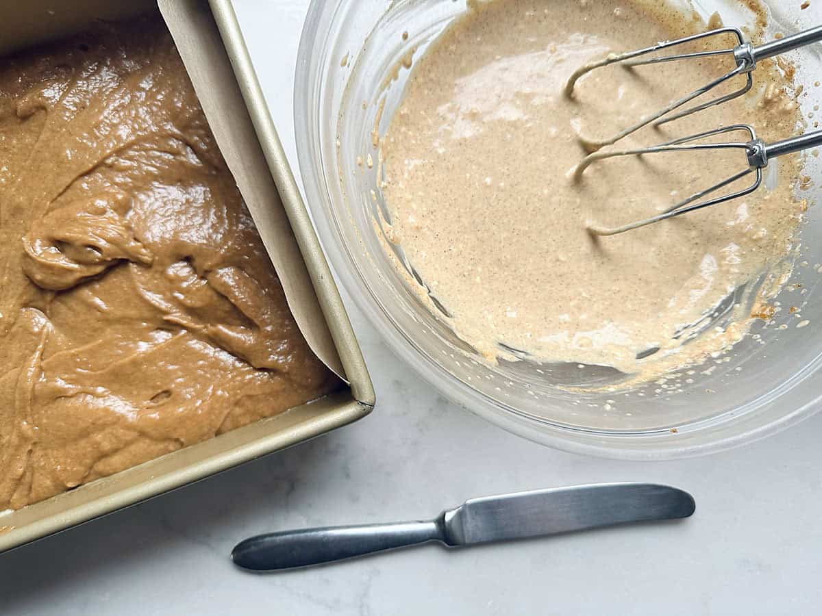 Cake batter in a rectangular baking pan next to a glass mixing bowl with an electric beater and a table knife.