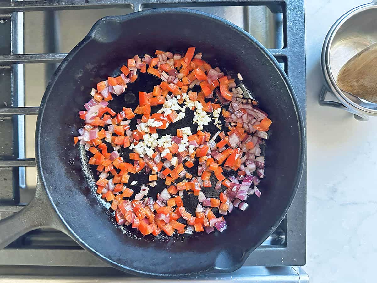 Diced red bell peppers, red onions, and minced garlic for a baked black-eyed pea dip cooking in a black cast iron skillet on a stovetop.