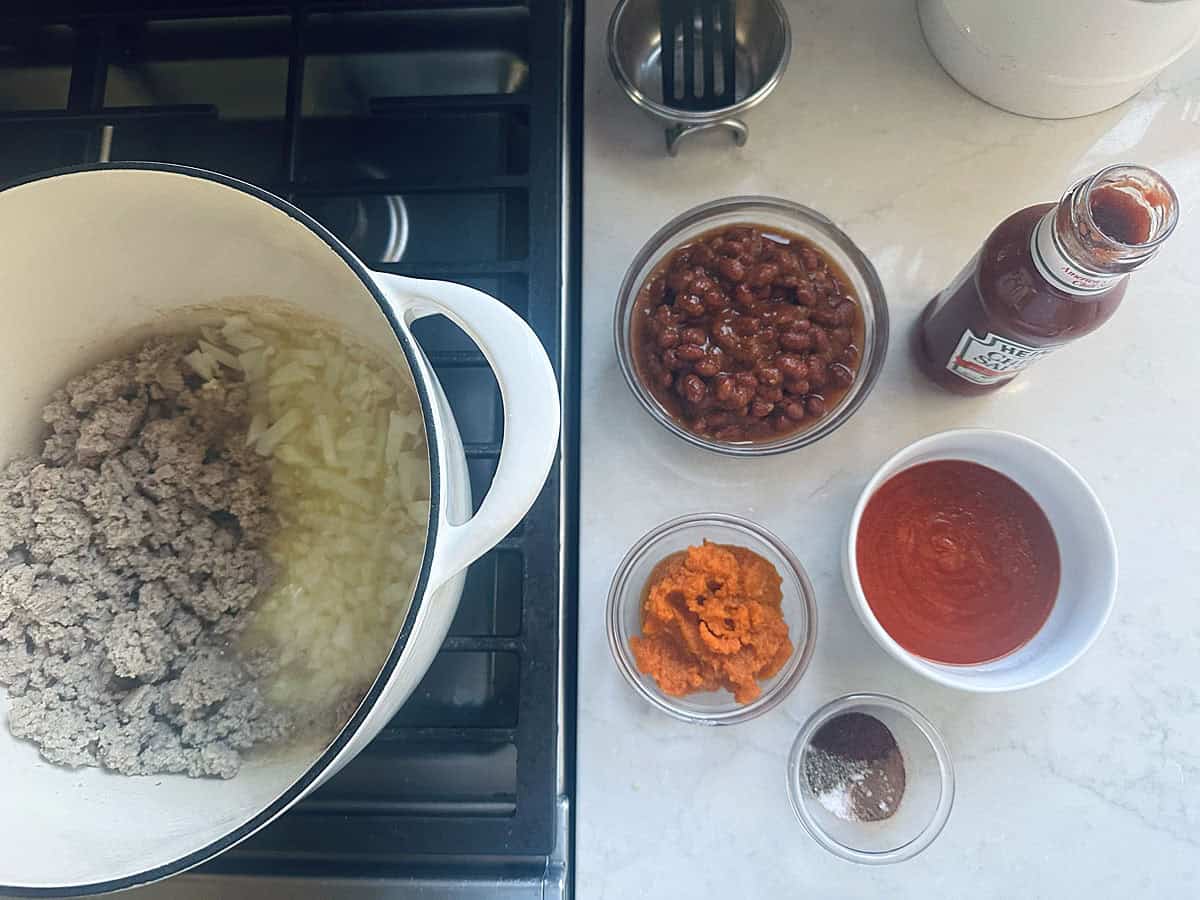 A large pot on a stove containing browned ground meat and chopped onions, alongside small bowls of beans, tomato sauce, pumpkin puree, spices, and a bottle of chili sauce.
