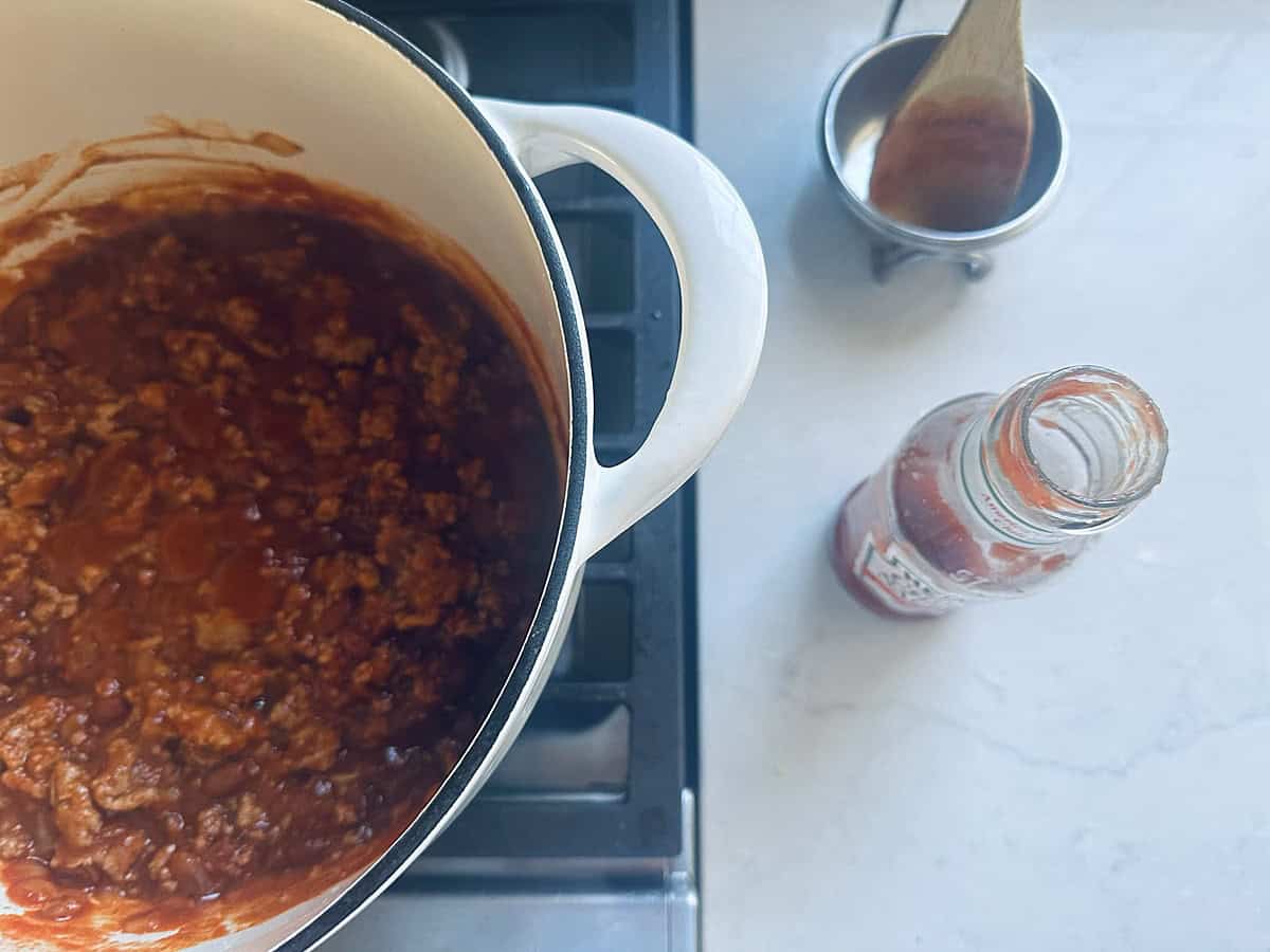 A white Dutch oven filled with meat sauce sits on a stovetop next to an empty glass sauce jar and a wooden spoon in a small metal dish.