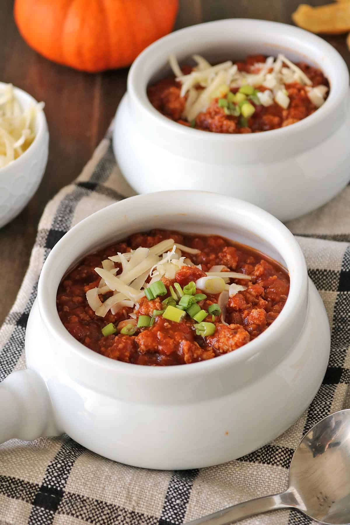 Two white bowls of turkey pumpkin chili topped with shredded cheese and sliced green onions, sitting on a plaid cloth.