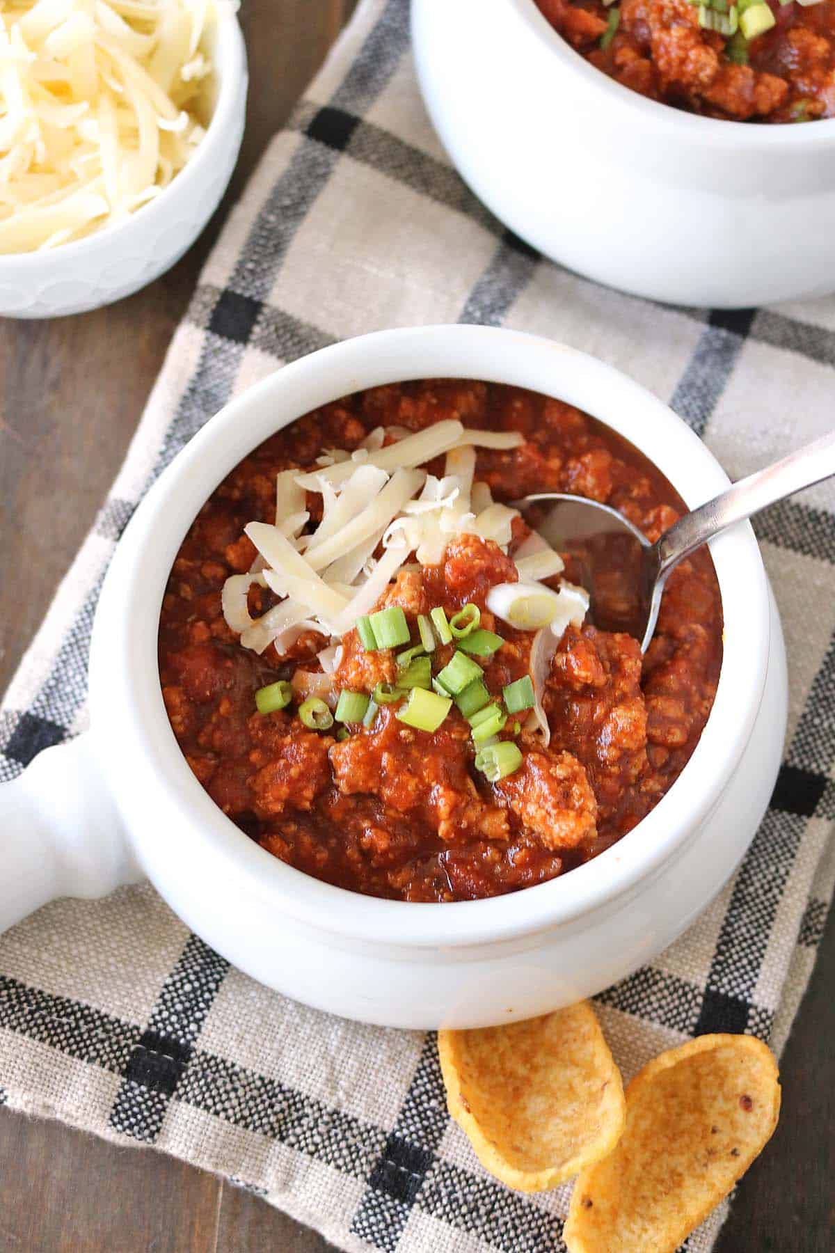 A bowl of turkey pumpkin chili topped with shredded cheese and green onions, served with a spoon and corn chips on a checkered napkin.