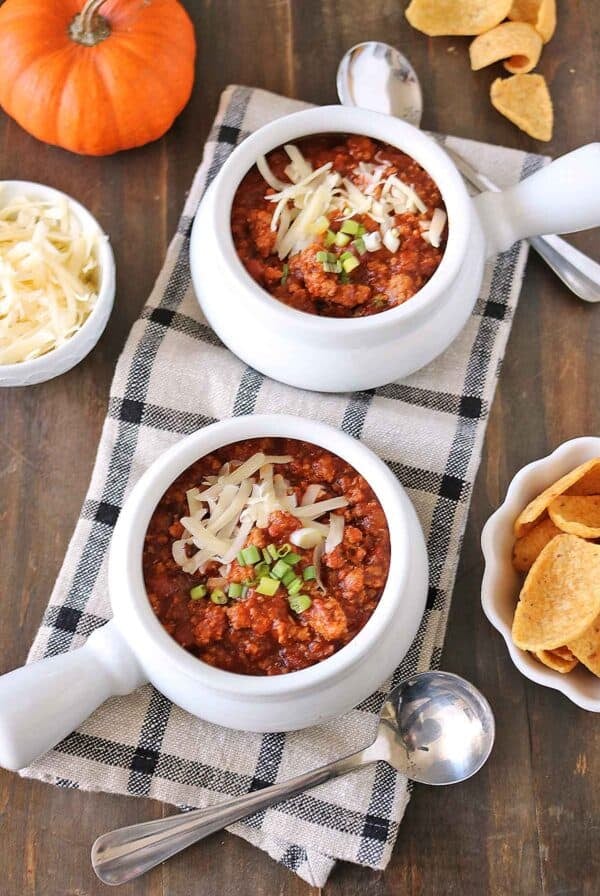 Two bowls of turkey pumpkin chili topped with shredded cheese and green onions, served with tortilla chips and a pumpkin nearby.